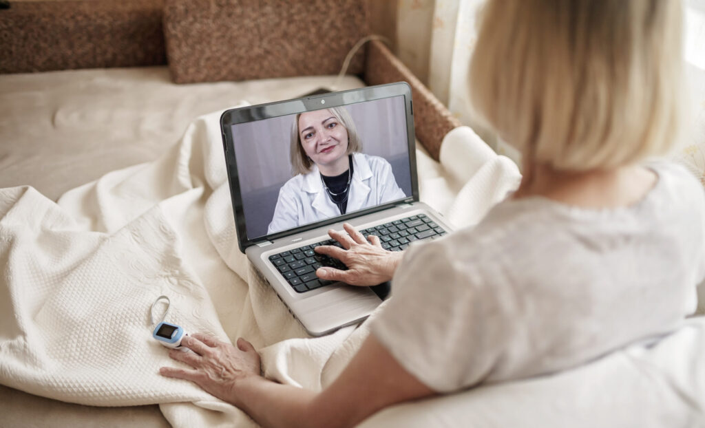 Old woman in bed looking at screen of laptop and consulting with a doctor online at home, telehealth